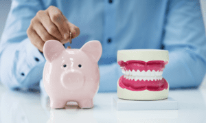 A hand placing a coin in a piggy bank, next to a model of human teeth, showing the need to improve dental cash flow with dental collection services.
