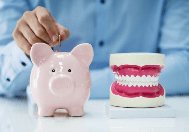 A hand placing a coin in a piggy bank, next to a model of human teeth, showing the need to improve dental cash flow with dental collection services.