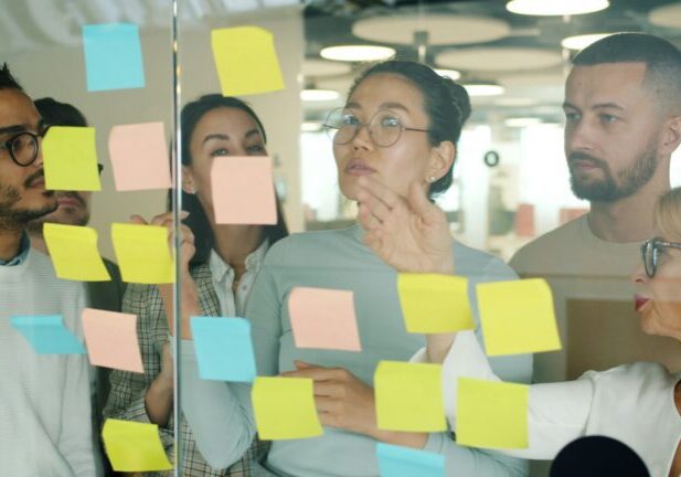 Group of colleagues discussing ideas on a glass wall covered with colorful sticky notes in a brainstorming session.