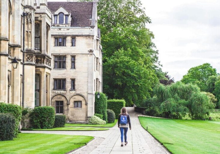 Person with a blue backpack walking along a stone path beside a historic beige building with Gothic windows and greenery on either side.